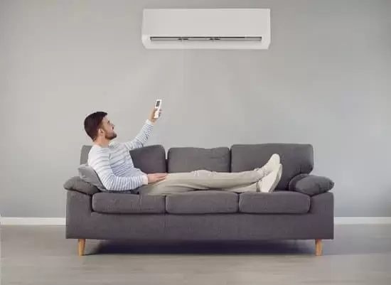 gentlema lying on a couch pointing remote control towards a wall mounted internal air conditioning unit